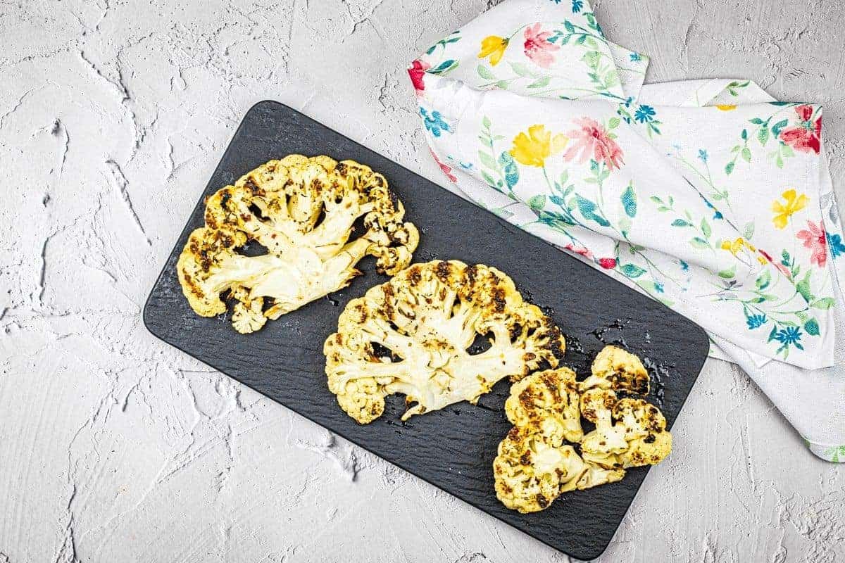 Three grilled cauliflower steaks on a black slate serving board with a floral napkin.