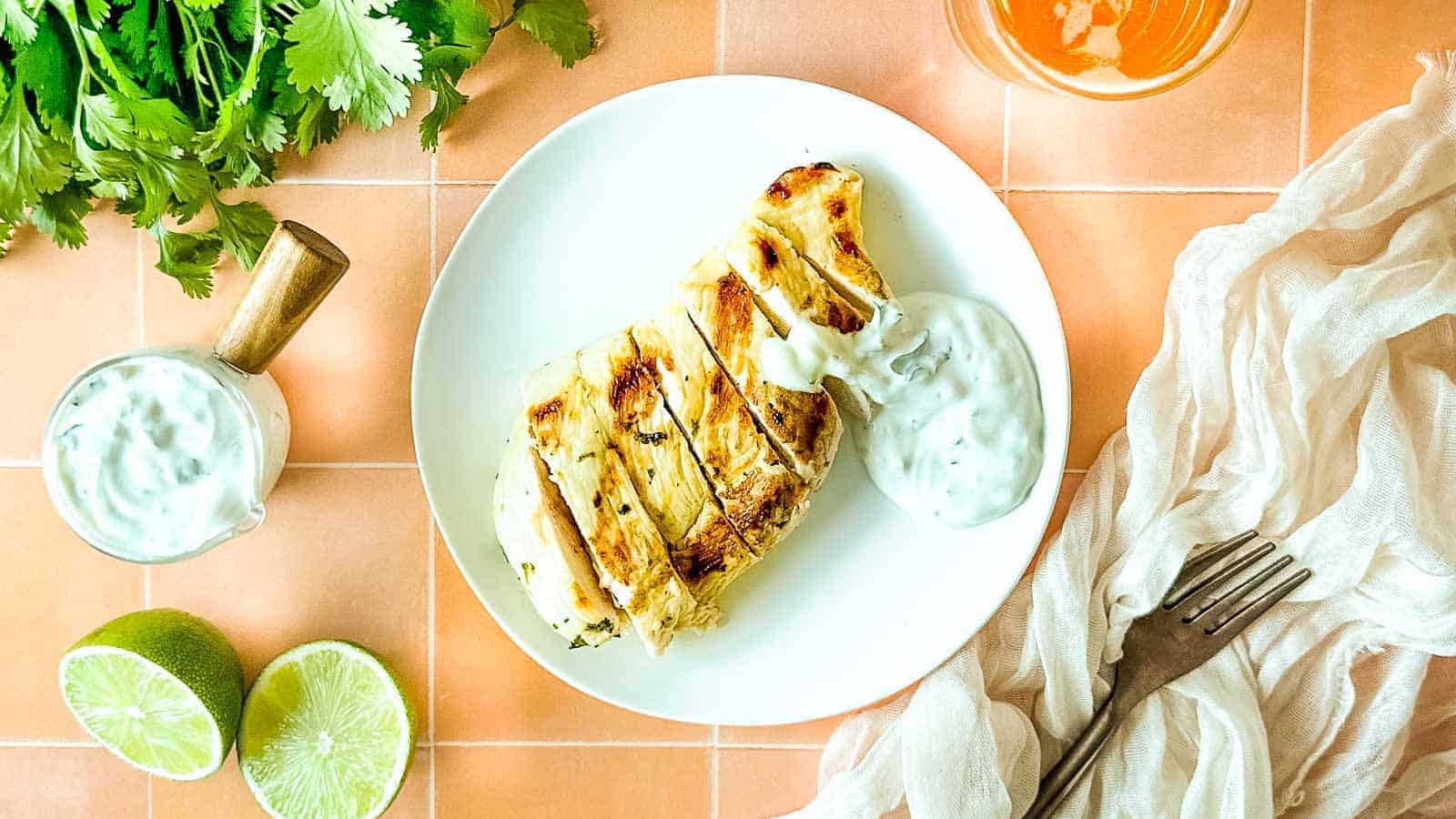 Sliced grilled chicken breast on a white plate with a dollop of cilantro crema and fresh lime halves nearby.