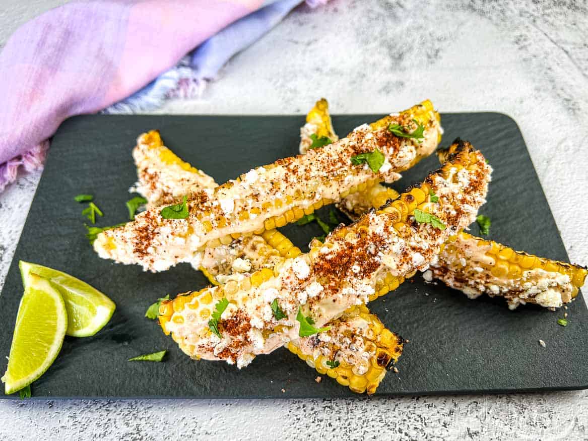 Grilled corn ribs topped with cheese, spices, and cilantro on a black slate board with lime wedges.