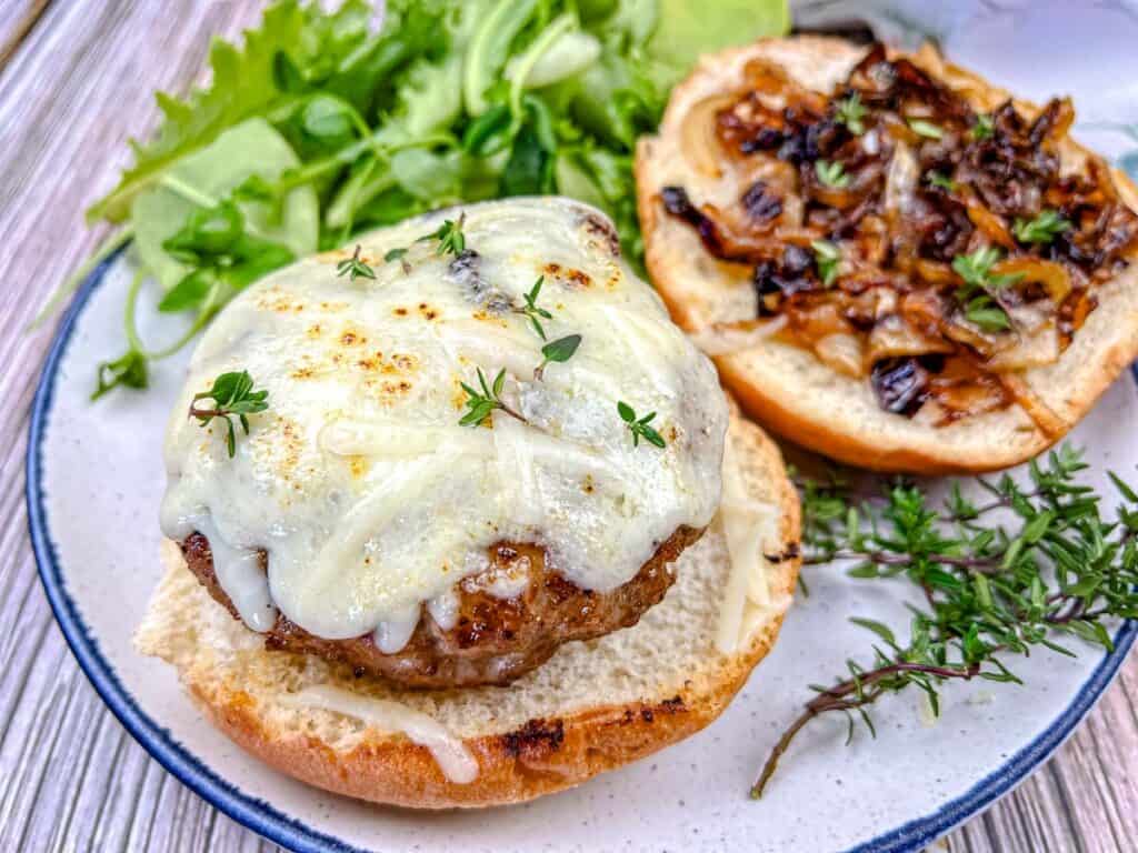Open-faced pork burger topped with melted cheese and caramelized onions, with greens on the side.
