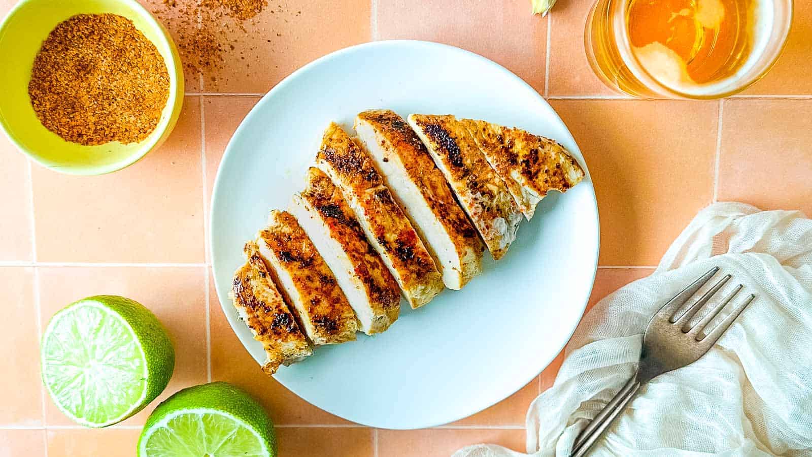 Sliced grilled chicken breast on a white plate with lime halves, seasoning, and a drink nearby.