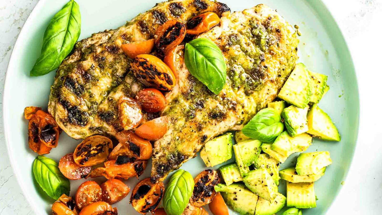 Grilled chicken breast with pesto, charred tomatoes, avocado chunks, and basil on a plate.
