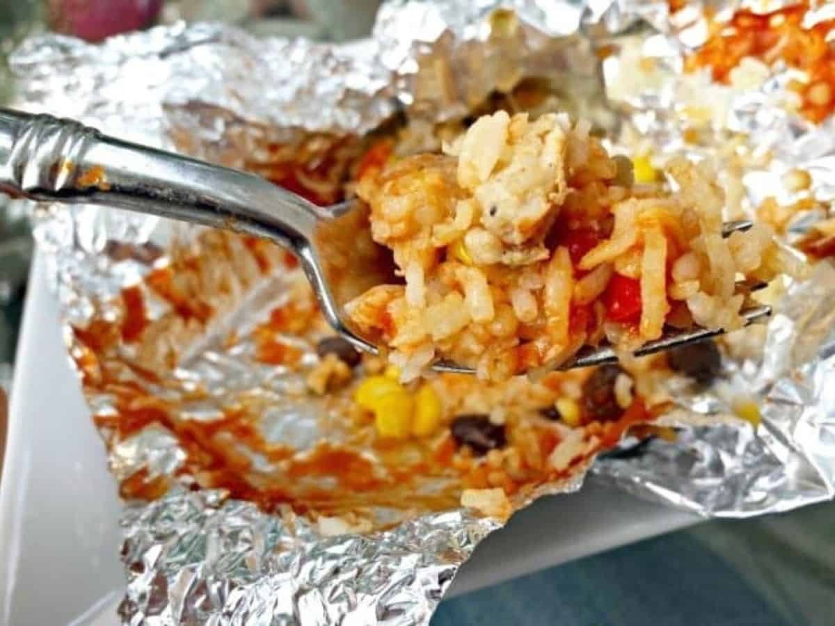 Fork holding a bite of cheesy rice, chicken, and corn over an open foil packet.